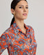 Model wearing the Sidney Paisley Liberty Silk Shirt in fiery orange-red—a 100% silk satin shirt featuring a paisley pattern, designed to elevate both country and city looks. Pair it with tailored trousers for a refined, office-ready ensemble, or dress down with jeans for a relaxed look. Front side view.