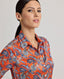 Model wearing the Sidney Paisley Liberty Silk Shirt in fiery orange-red—a 100% silk satin shirt featuring a paisley pattern, designed to elevate both country and city looks. Pair it with tailored trousers for a refined, office-ready ensemble, or dress down with jeans for a relaxed look. Front side view.