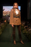 A model walks a fashion runway wearing a brown double-breasted cape jacket with a white ruffled collar, brown pants, and tall brown boots. The runway is lined with greenery, and a large screen in the background displays a landscape scene.