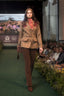 A model walks the runway wearing a brown tweed blazer with brown corduroy trousers and a red patterned scarf. The runway has a green carpet, with an audience seated on both sides and a screen in the background showing a landscape and the text 'Kings Trust 50'.
