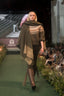 A model walks down a fashion runway wearing a green knit sweater with a fairilse pattern, a dark green fringed shawl, dark green pants, and tall brown leather boots. The runway is green with an audience seated on both sides.