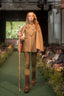 A model walks a fashion runway wearing a tan double-breasted coat with a matching cape, brown pants, and tall brown boots, while holding a wooden staff. The runway is lined with guests, and a green screen in the background displays a forest scene.