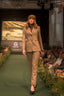 A model walks down a fashion runway wearing a beige plaid tailored suit with a matching patterned hat. The runway is green with seated spectators on both sides, and a screen in the background shows a landscape image with the text 'Kings Trust'.