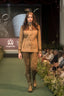 A model walks a fashion runway wearing a tan and brown jacket with matching brown trousers and tall brown boots. The runway is green with seated audience members on both sides, and a King's Trust banner is visible in the background.