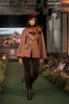 A model walks the runway wearing a tan cape jacket with a floral accent, dark brown pants, and a burgundy beret. The runway is green carpeted with an audience seated on both sides.