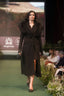 A model walks the runway wearing a long black coat with a large dark fur collar, holding a black clutch. The runway has a green carpet, with seated audience members on the right and a King's Trust banner in the background.