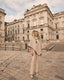 Woman in a beige cape outfit standing in front of a large building with classical architecture.