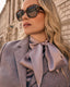 Woman wearing sunglasses and a purple safari jacket with a silk blouse, standing against a stone building.