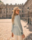Woman in a light blue blazer and beige fedora standing on a cobbled street with classical architecture in the background.