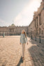 Woman in a light blue safari jacket and hat walking in an open courtyard with classical architecture.