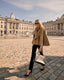 Woman in a brown trench cape standing in an urban setting with classical architecture.