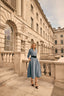 Woman in a blue dress standing in front of an elegant building with classical architecture.