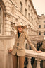 Woman in a beige safari jacket and hat standing in front of an elegant building.