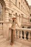 Woman in a beige safari jacket and brown trousers standing on a stone balcony with classical architecture in the background