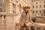 Woman in a beige safari jacket and hat standing in front of an architectural building.