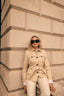 Woman in a beige trench coat and sunglasses leaning against a stone wall.