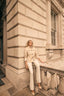 Woman in a beige trench coat and white pants sitting on a stone ledge of a classical building.