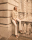Woman in a beige trench coat and white pants sitting on a stone ledge of an old building.