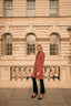Woman in a pink coat standing in front of a stone building with arched windows.