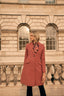 Woman in a pink coat standing in front of a stone building with arched windows.