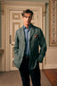 A man wearing a textured sage green blazer over a light blue button-down shirt and dark trousers, standing in an elegant indoor setting with a decorative door behind him.