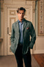 A man wearing a textured sage green blazer over a light blue button-down shirt and dark trousers, standing in an elegant indoor setting with a decorative door behind him.