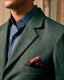 Close-up of a man wearing a textured green herringbone blazer over a blue button-down shirt, with a patterned pink pocket square in the breast pocket.