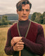 A man wearing a burgundy and forest green knit sweater vest over a dark burgundy turtleneck sweater, standing in a grassy field with trees and hills in the background.