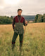 A man stands in a grassy field holding a walking stick, wearing a vintage-style hunting outfit with a maroon and gray-green cardigan, matching trousers, tall boots, and a dark turtleneck. Trees, hills, and a bridge are visible in the background under an overcast sky.