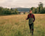 A man stands in a grassy field holding a walking staff, wearing a vintage-style hunting outfit with a maroon and green jacket, dark trousers, and tall rubber boots, with a bridge and trees in the background.