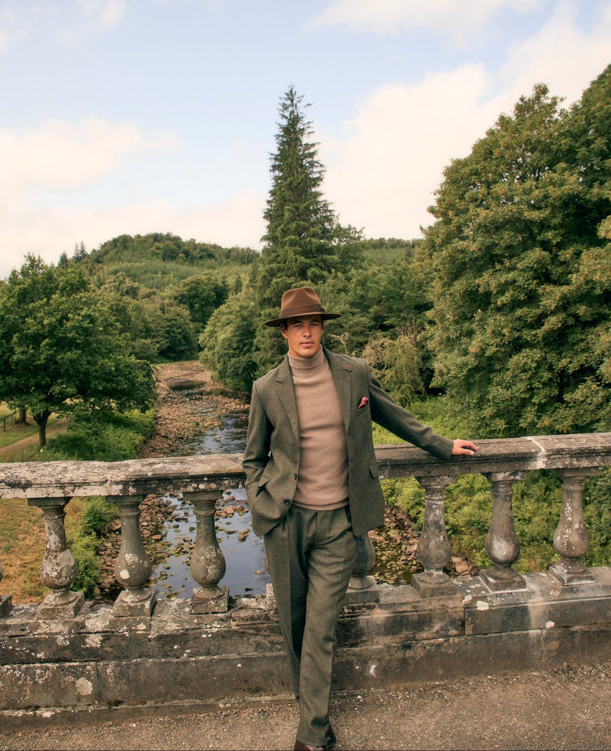 A man wearing a brown fedora, olive green tweed suit, beige turtleneck, and brown leather shoes stands on a stone bridge in a scenic outdoor setting with trees and a river in the background.