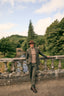 A man wearing a brown fedora, olive green tweed suit, beige turtleneck, and brown leather shoes stands on a stone bridge in a scenic outdoor setting with trees and a river in the background.