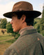 A man in profile wearing a brown wide-brim fedora hat, a beige turtleneck, and a brown tweed jacket with a subtle red windowpane pattern, set against a blurred outdoor park background.