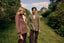 A man and a woman stand on a grassy path in front of a castle, wearing coordinated tweed and wool-style outerwear. The woman wears a dusty mauve-brown jacket with a dark burgundy yoke and white scarf, while the man wears a taupe-brown blazer with dark plum trousers.