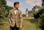 A man wearing a brown tweed blazer with a patterned pocket square stands in a grassy field with a castle in the background under a cloudy sky.