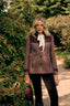 A woman stands outdoors on a garden path wearing a color-blocked tweed jacket in dusty rose and dark burgundy with a cream bow tie, paired with black pants and holding a matching burgundy hat.