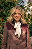 A woman with blonde hair wearing a brown and burgundy color-blocked coat with a cream ribbon tie, standing outdoors in front of green foliage.