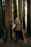 A woman stands in a forest between large tree trunks, wearing a brown double-breasted cape-style coat over a white top, black pants, and tall black boots, while holding a wooden walking stick.
