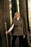 A woman stands in a forest between large tree trunks, wearing a brown double-breasted cape-style coat with a dark brown belt, a white turtleneck, black pants, and holding a wooden staff.
