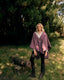 A woman standing outdoors in a grassy area wearing a mauve-purple wrap poncho over a cream collared shirt, with dark pants and holding a pair of dark gloves.