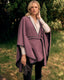 A woman standing outdoors in a grassy area wearing a mauve-purple wrap poncho over a cream collared shirt, with dark pants and holding a pair of dark gloves.