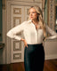 A woman stands in an elegant interior room with wood floors and paneled doors, wearing a fitted white long-sleeve blouse and a dark navy mermaid-style skirt with a train.