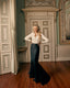 A woman stands in an elegant interior room with wood floors and paneled doors, wearing a fitted white long-sleeve blouse and a dark navy mermaid-style skirt with a train.