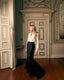 A woman stands in an elegant interior room with wood floors and paneled doors, wearing a fitted white long-sleeve blouse and a dark navy mermaid-style skirt with a train.