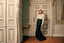 A woman stands in an elegant room with paneled doors and wood flooring, wearing a white long-sleeve mock-neck top and a dark navy satin mermaid-style skirt.