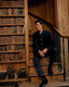 A man in a dark navy military-style jacket and tartan plaid trousers sits on wooden stairs next to a large bookshelf filled with vintage books.
