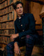 A man wearing a dark navy jacket and purple plaid trousers sits on wooden stairs in front of a bookshelf.