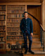A man stands on wooden stairs in a vintage library, wearing a dark navy blue jacket with red piping and matching plaid trousers.