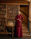 A woman stands in a grand library wearing a long, deep burgundy satin-like wrap dress. She is posed next to a vintage globe on a wooden table, with tall bookshelves and a wooden staircase behind her.