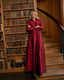 A woman stands on wooden stairs in a library, wearing a long-sleeved, wrap-style maxi dress in deep burgundy satin. She has her arms crossed, with bookshelves and a wooden staircase behind her.